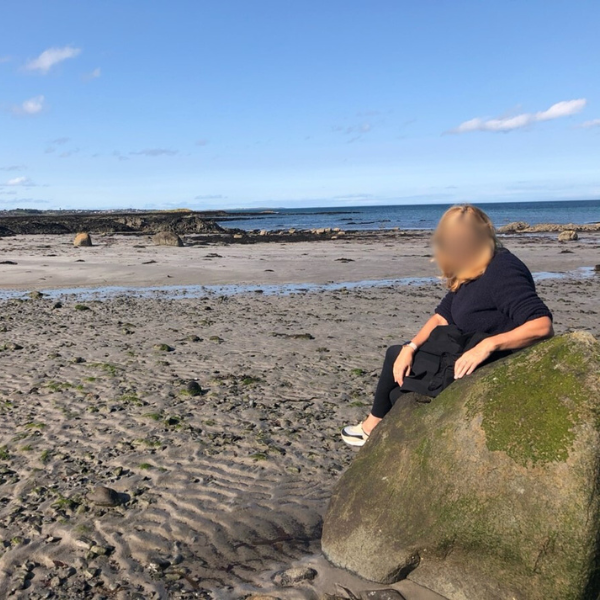 Photo of a woman with her face blurred, sitting on some cliffs next to the beach, dressed in black